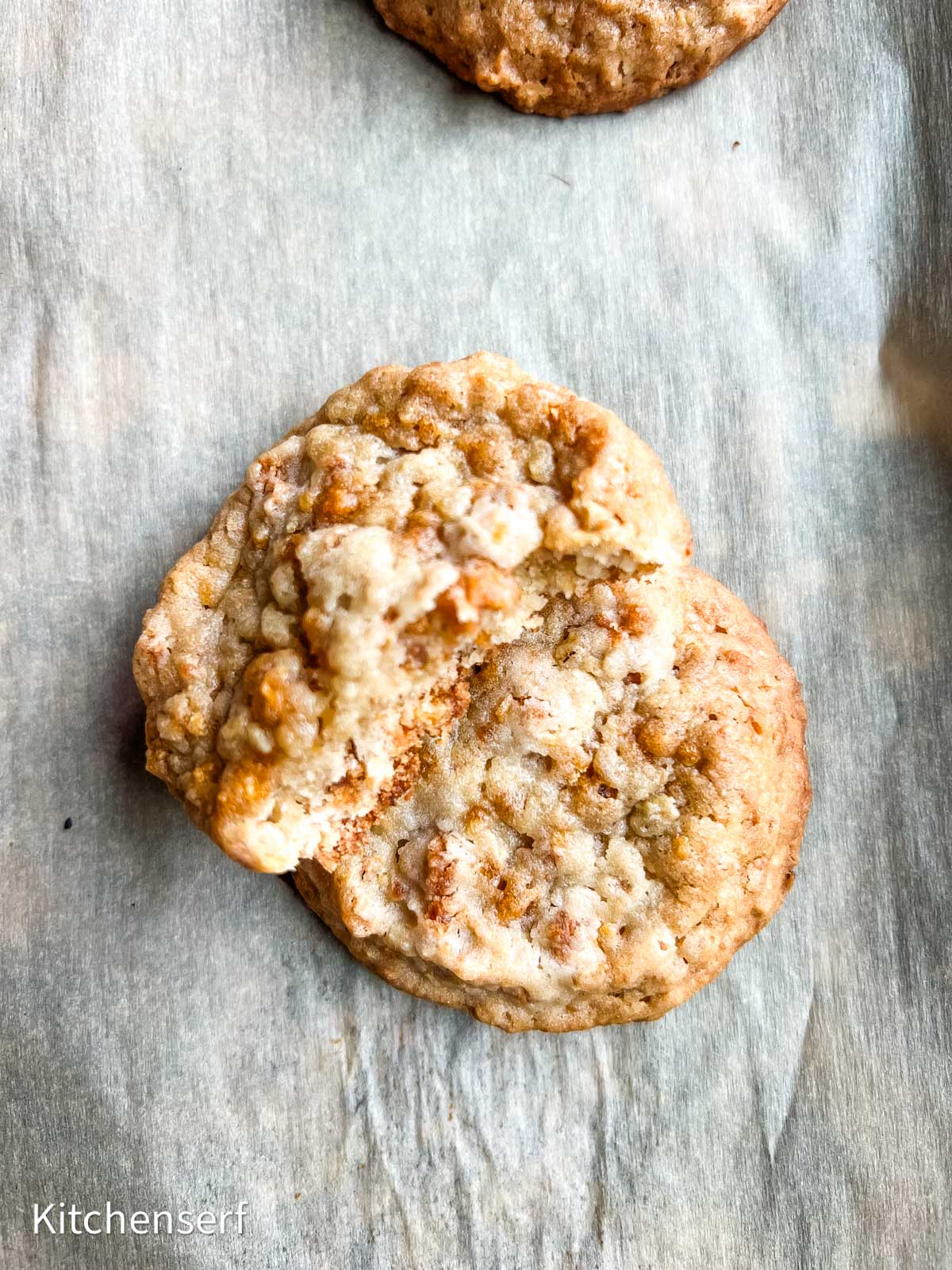 Two oatmeal cookies on parchment paper, one with a bite taken out of it.