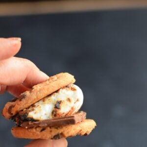 A hand holding a smore made with chocolate chip cookies, melted chocolate, and a toasted marshmallow.