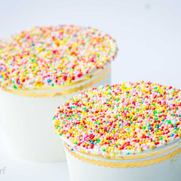 Two white cakes topped with colorful round sprinkles, inspired by the viral dot cake recipe, and trimmed with a gold scalloped border on a white background.