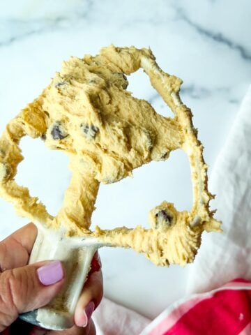 Hand holds a mixer paddle covered in chocolate chip cookie dough, showcasing the chocolate chip cookie dough texture against a marble background.