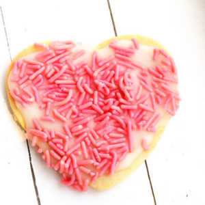 Heart-shaped cookie with white icing and pink sprinkles on a white wooden surface.