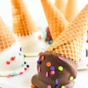 Waffle cones dipped in chocolate and white coatings, topped with colorful sprinkles, on a white surface.