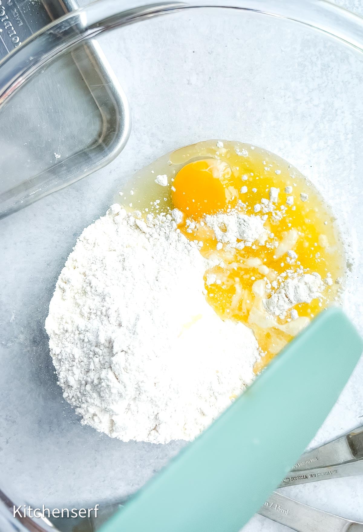 Glass bowl with fcake mix, and cracked eggs, a green spatula, and metal pan on a light countertop—perfect for whipping up half batch cake mix cookies.