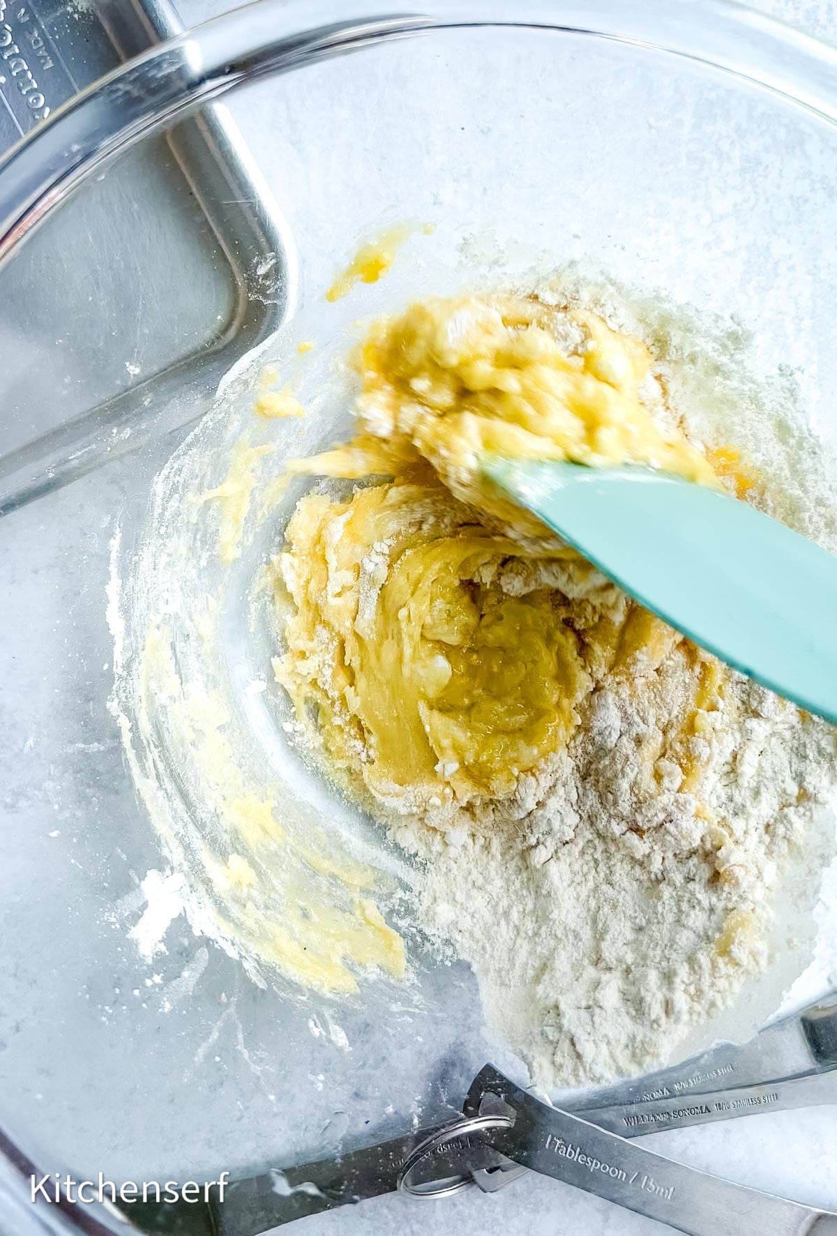 A blue spatula mixes dough with flour in a clear glass bowl; measuring spoons are nearby, ready for making half batch cake mix cookies.
