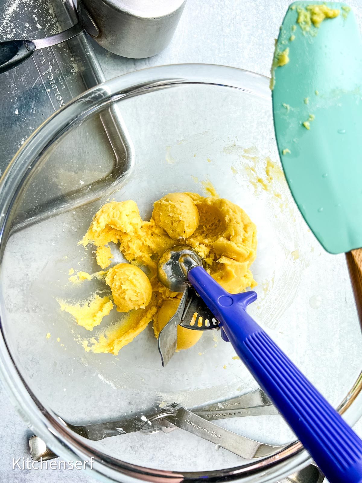 Cookie dough for half batch cake mix cookies with a scoop in a glass bowl, spatula and utensils nearby on a light surface.