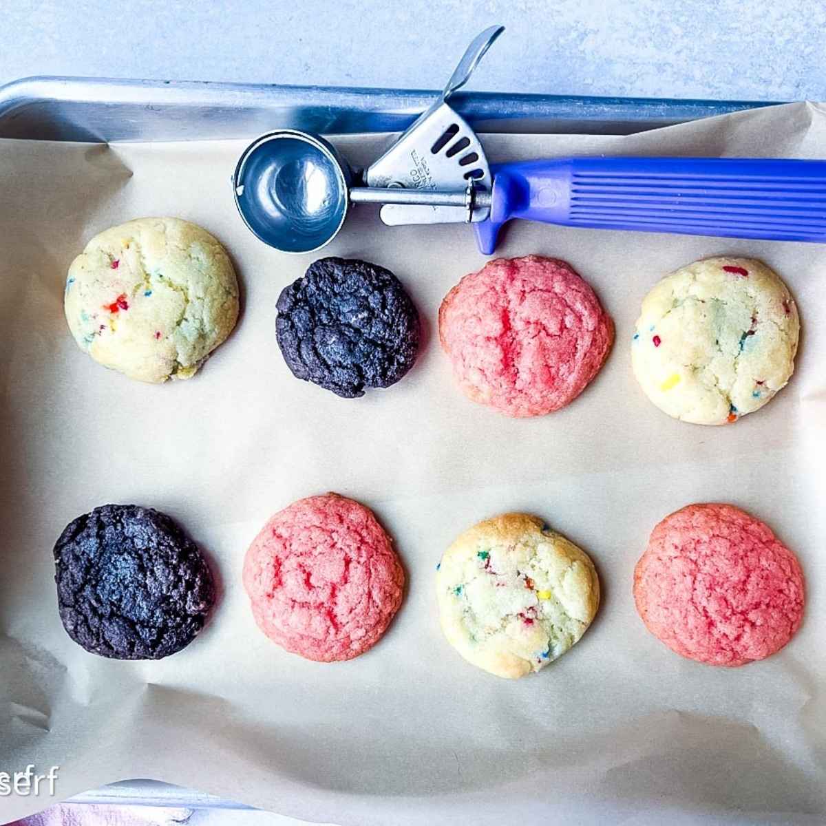 A baking tray with eight colorful half batch cake mix cookies and a purple cookie scoop on parchment paper.