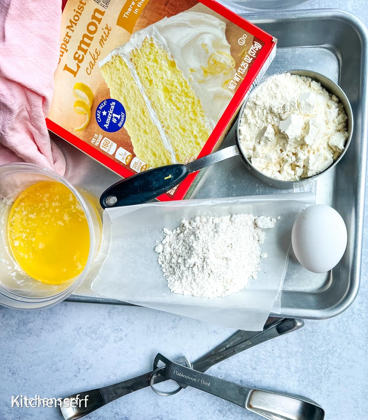 Cake mix, flour, egg, melted butter, measuring spoons, and a pink towel on a metal tray—everything you need to make delicious half batch cake mix cookies.
