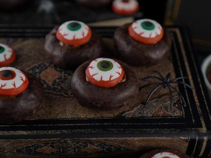 Halloween Eyeball Donuts: Chocolate donuts decorated with candy eyeballs rest on an old book, with a fake spider nearby. A perfectly spooky Halloween treat.