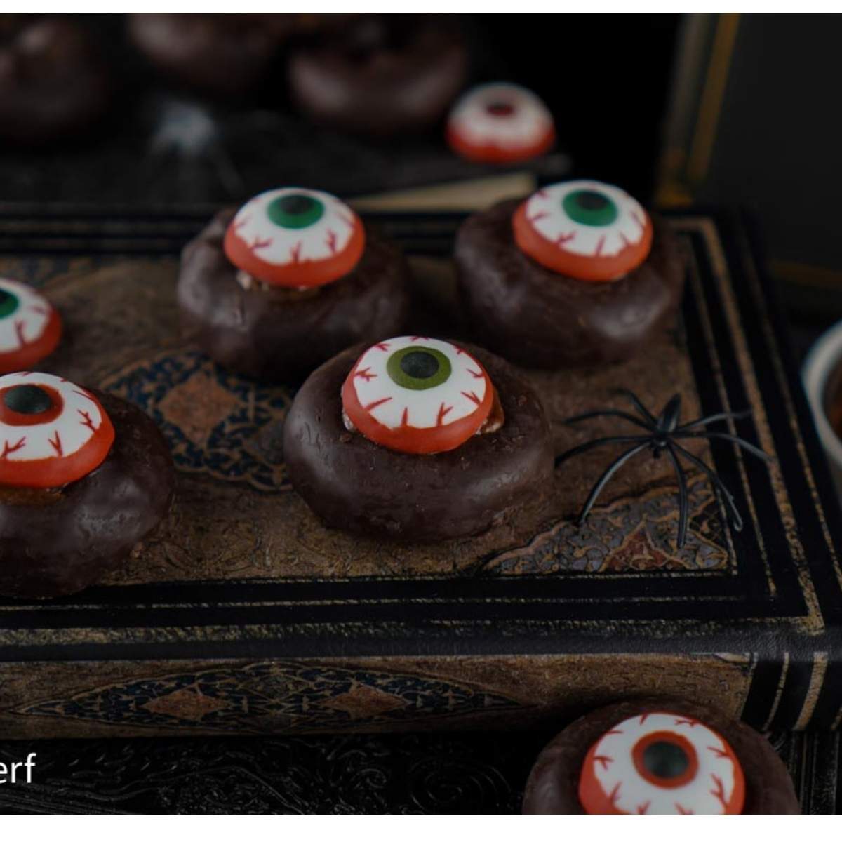 Halloween Eyeball Donuts: Chocolate donuts decorated with candy eyeballs rest on an old book, with a fake spider nearby. A perfectly spooky Halloween treat.