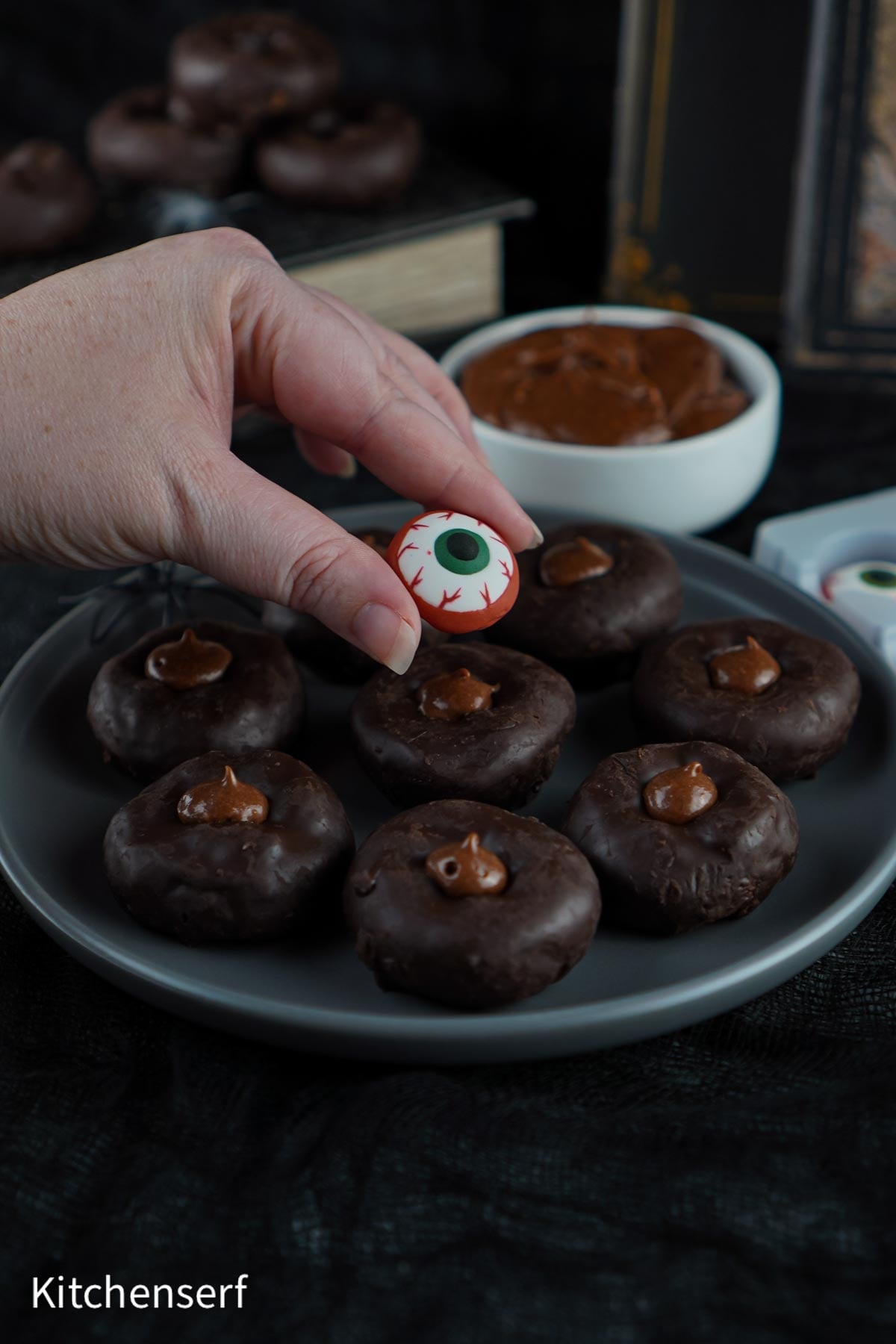 A hand holds a candy eyeball over a plate of chocolate-covered treats, creating the perfect Halloween Eyeball Donuts scene, with a bowl of chocolate in the background.