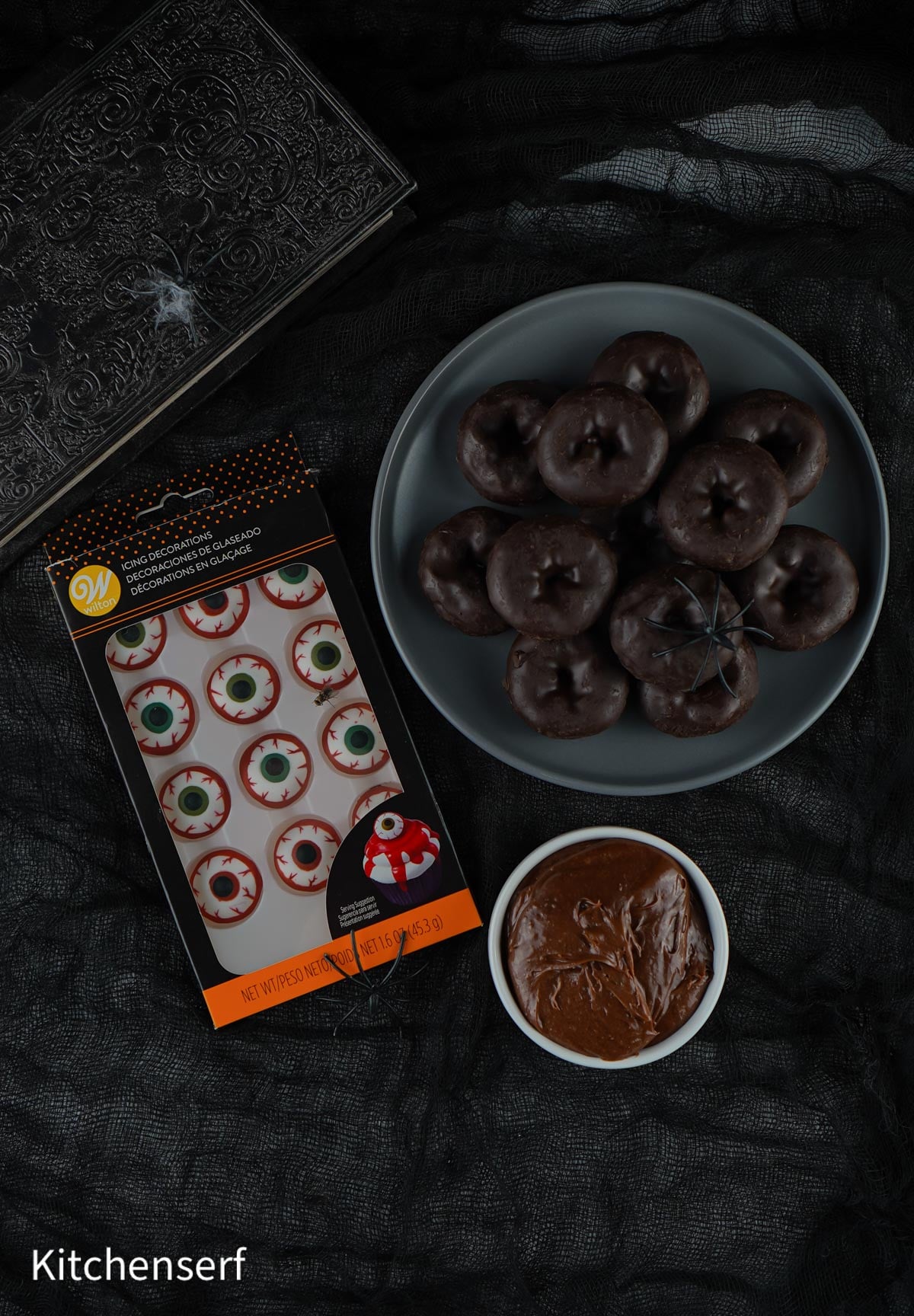 A plate of Halloween Eyeball Donuts, candy eyeballs, and a bowl of chocolate spread on a dark cloth background.