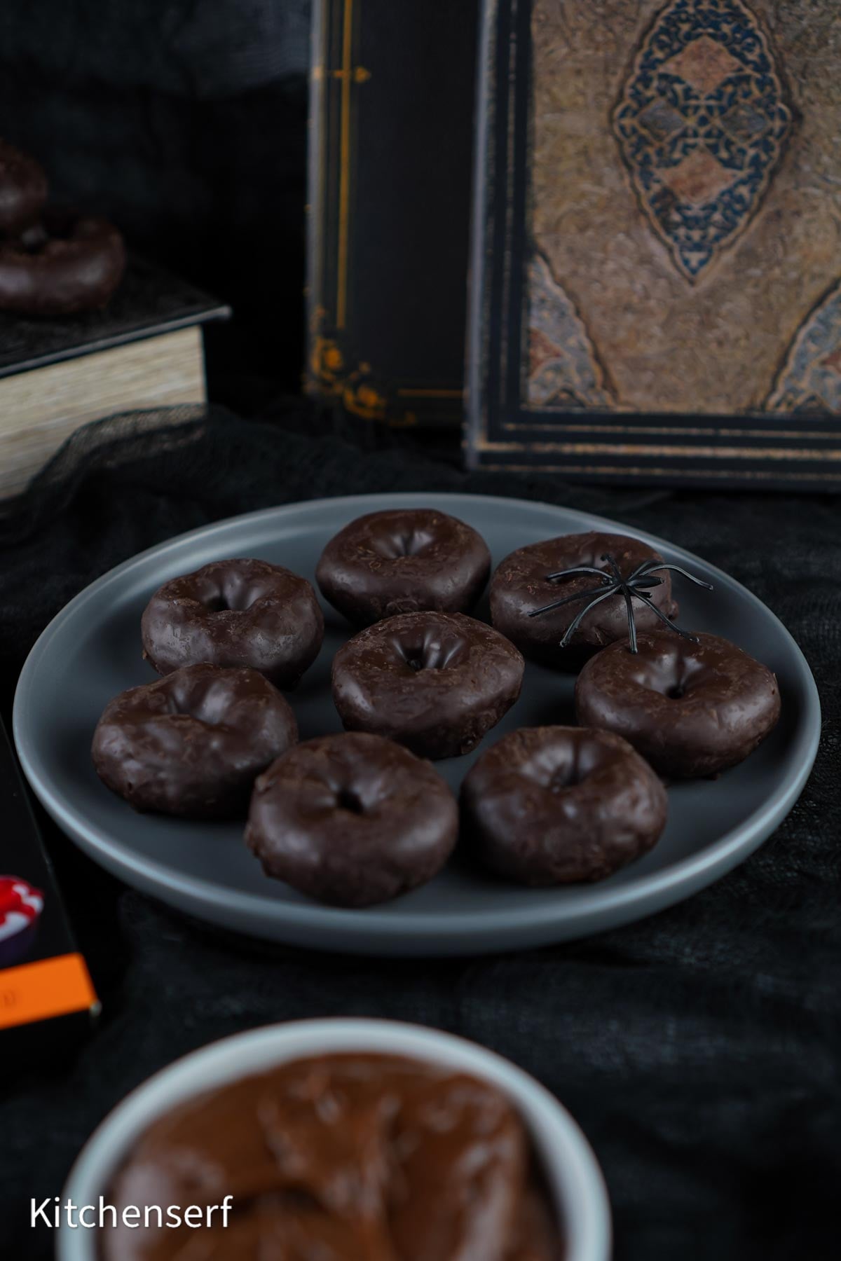 Chocolate mini donuts on a gray plate with a plastic spider, surrounded by books and a bowl of chocolate, make the perfect Halloween Eyeball Donuts display for your spooky celebration.