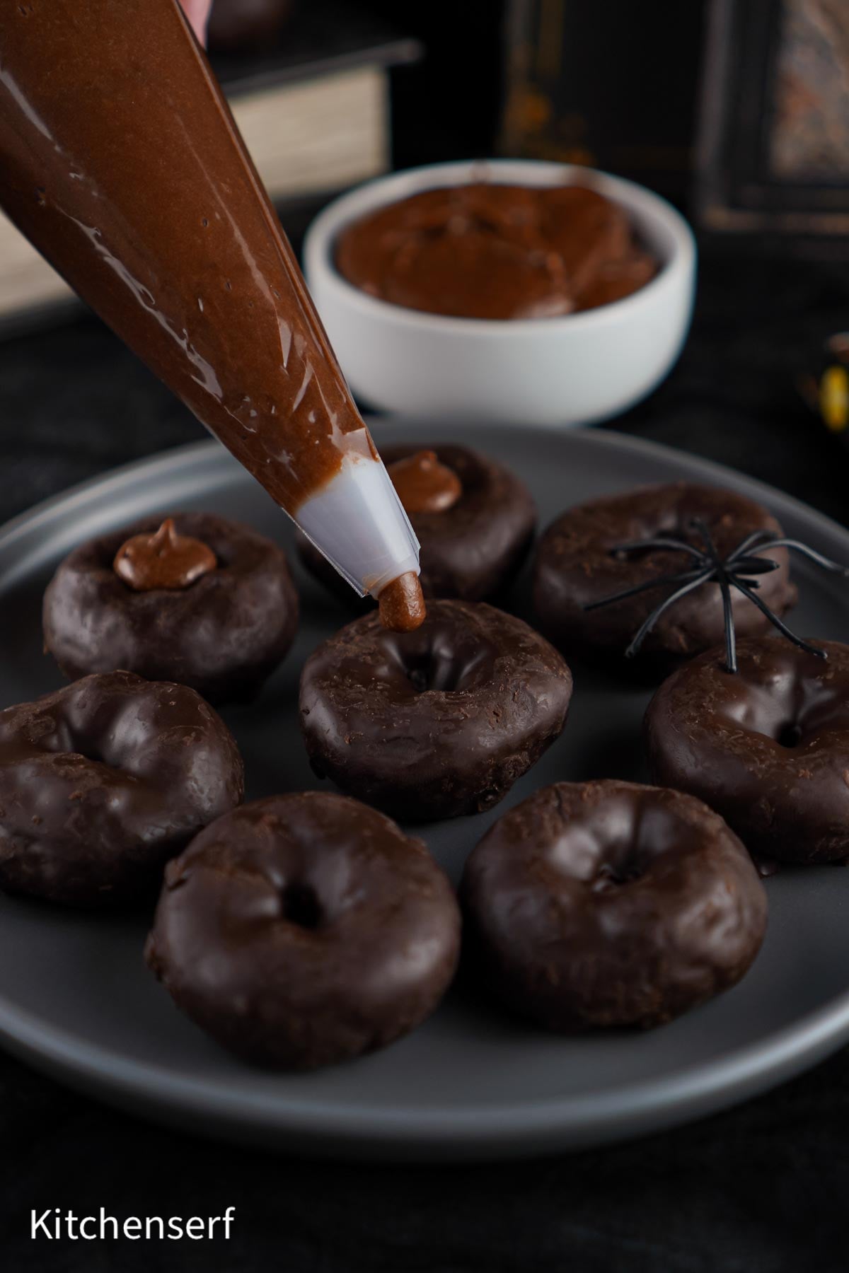 A piping bag fills mini chocolate donuts; a bowl of chocolate and a fake spider are in the background, creating the perfect spooky setup for Halloween Eyeball Donuts.