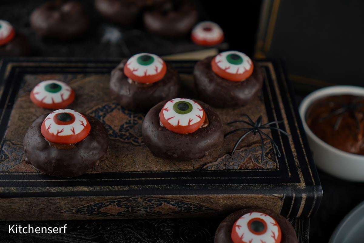 Halloween Eyeball Donuts are chocolate donuts topped with candy eyeballs, arranged on an old book with a plastic spider and more treats nearby.
