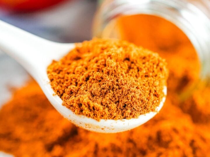A white spoon holding ground cinnamon with more cinnamon spilled from a glass jar in the background.