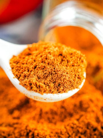 A white spoon holding ground cinnamon, with more fresh cinnamon spilled from a jar in the background, evokes a scene similar to learning how to make ground cinnamon—highlighting the vibrant color and freshness of homemade spices.