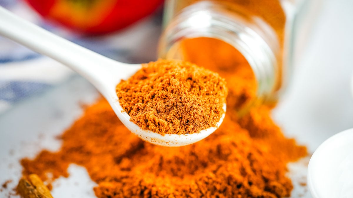 A white spoon holding ground cinnamon, with more fresh cinnamon spilled from a jar in the background, evokes a scene similar to learning how to make ground cinnamon—highlighting the vibrant color and freshness of homemade spices.