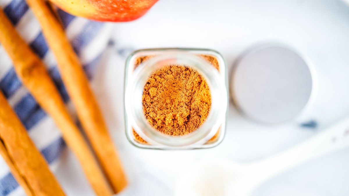 Top view of a jar of ground cinnamon, cinnamon sticks, an apple, and a spoon on a white surface—perfect for illustrating how to make ground cinnamon at home.