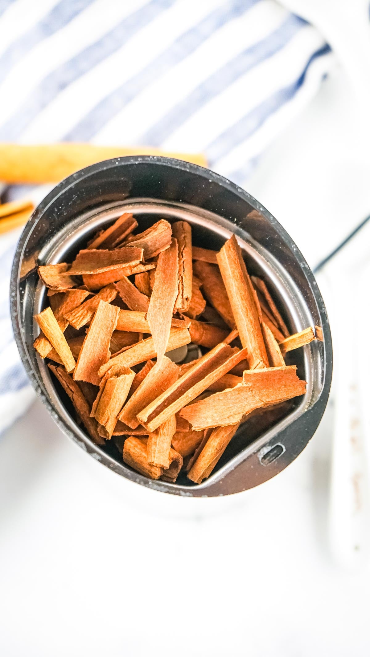 A metal measuring cup filled with cinnamon sticks rests on a white surface with a striped cloth nearby, perfect for illustrating how to make ground cinnamon at home.