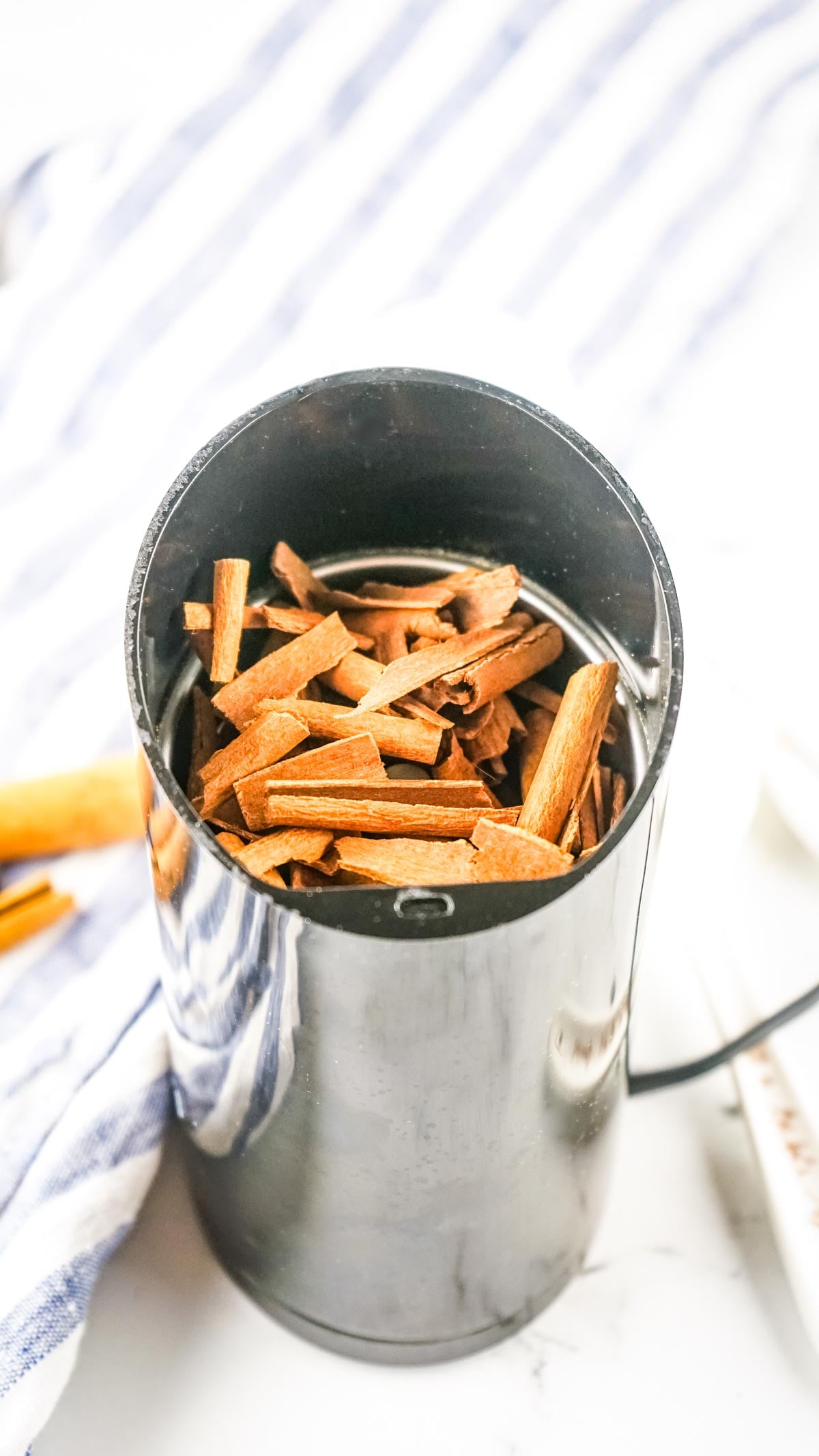 A black electric grinder filled with pieces of cinnamon sticks on a white surface with a striped cloth nearby, perfectly illustrating how to make ground cinnamon at home.