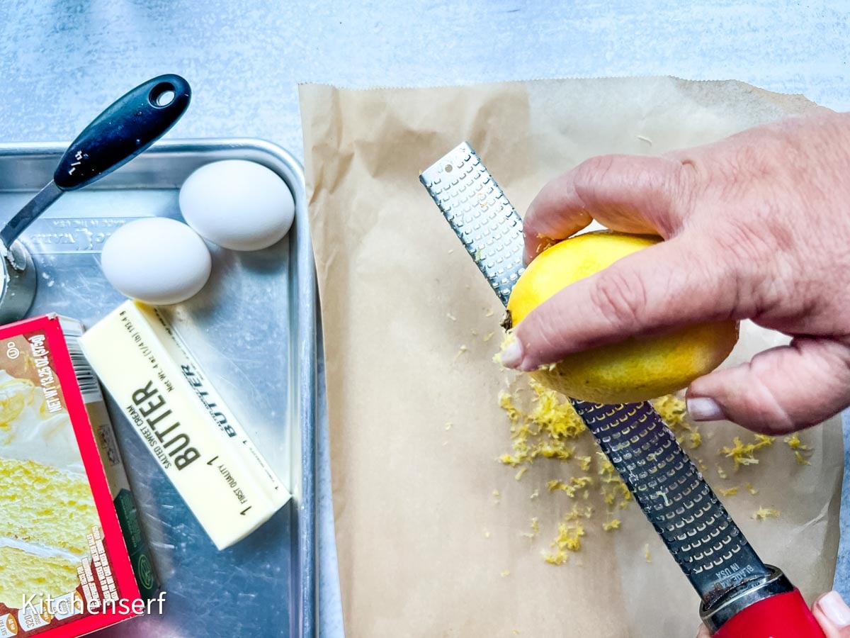 A hand zesting a lemon over parchment paper, with eggs, butter, and utensils nearby on a tray—perfect prep for lemon cake mix cookies.