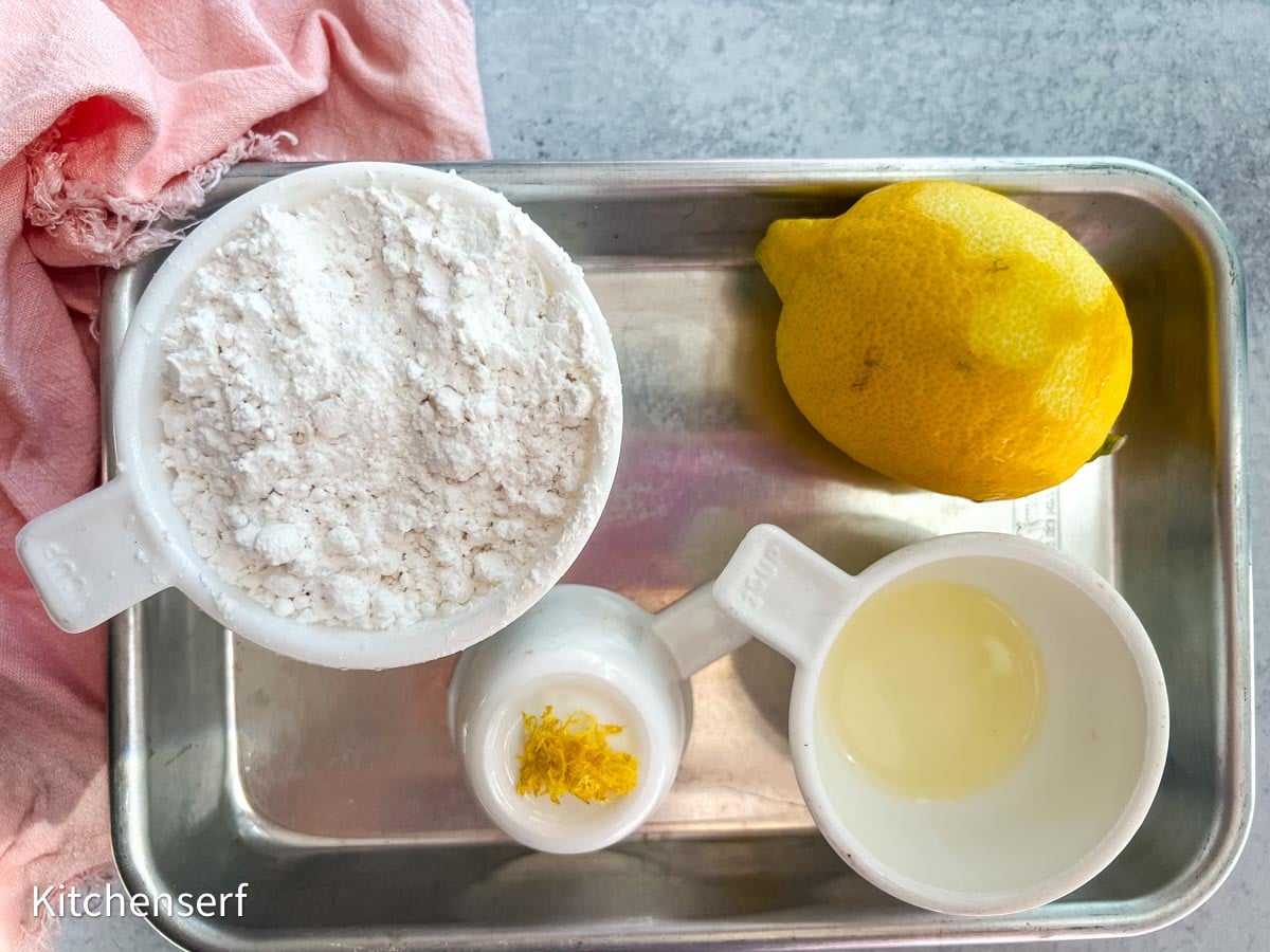A tray with flour, a lemon, lemon juice, and lemon zest in separate cups—perfect ingredients for lemon cake mix cookies—rests beside a pink cloth.