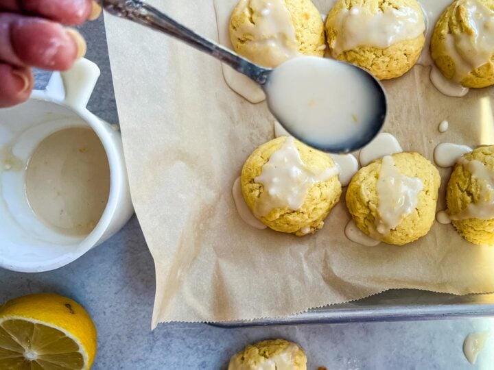 Hand drizzling glaze over lemon cake mix cookies on a parchment-lined tray, with fresh lemon and a bowl of glaze nearby.