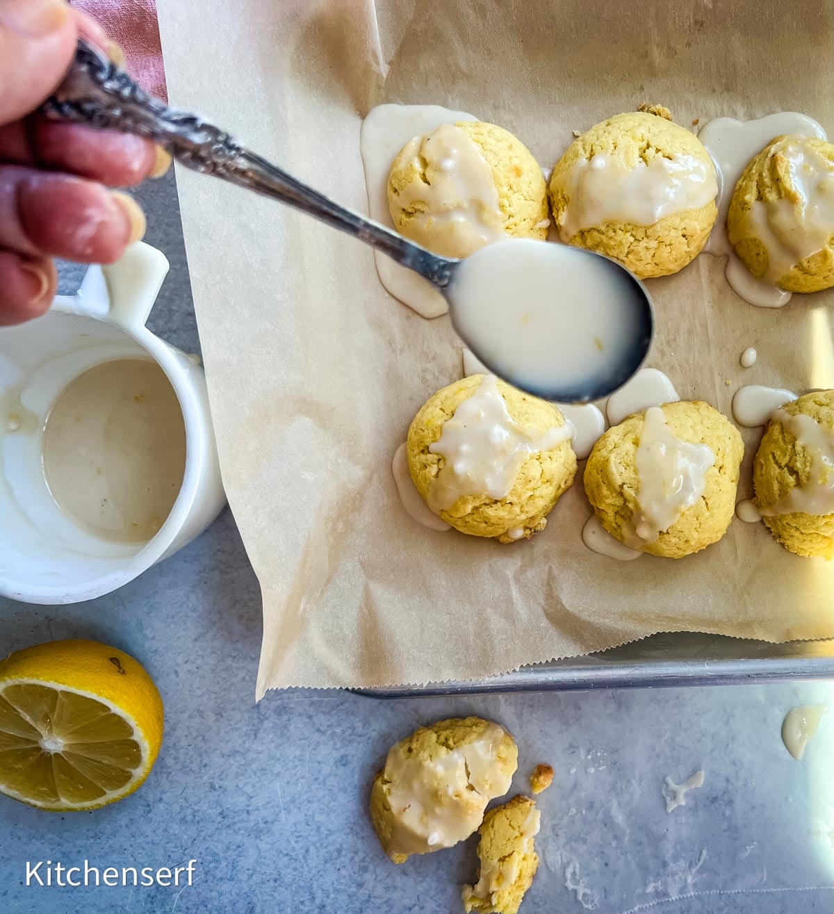 Hand drizzling glaze over lemon cake mix cookies on a parchment-lined tray, with fresh lemon and a bowl of glaze nearby.