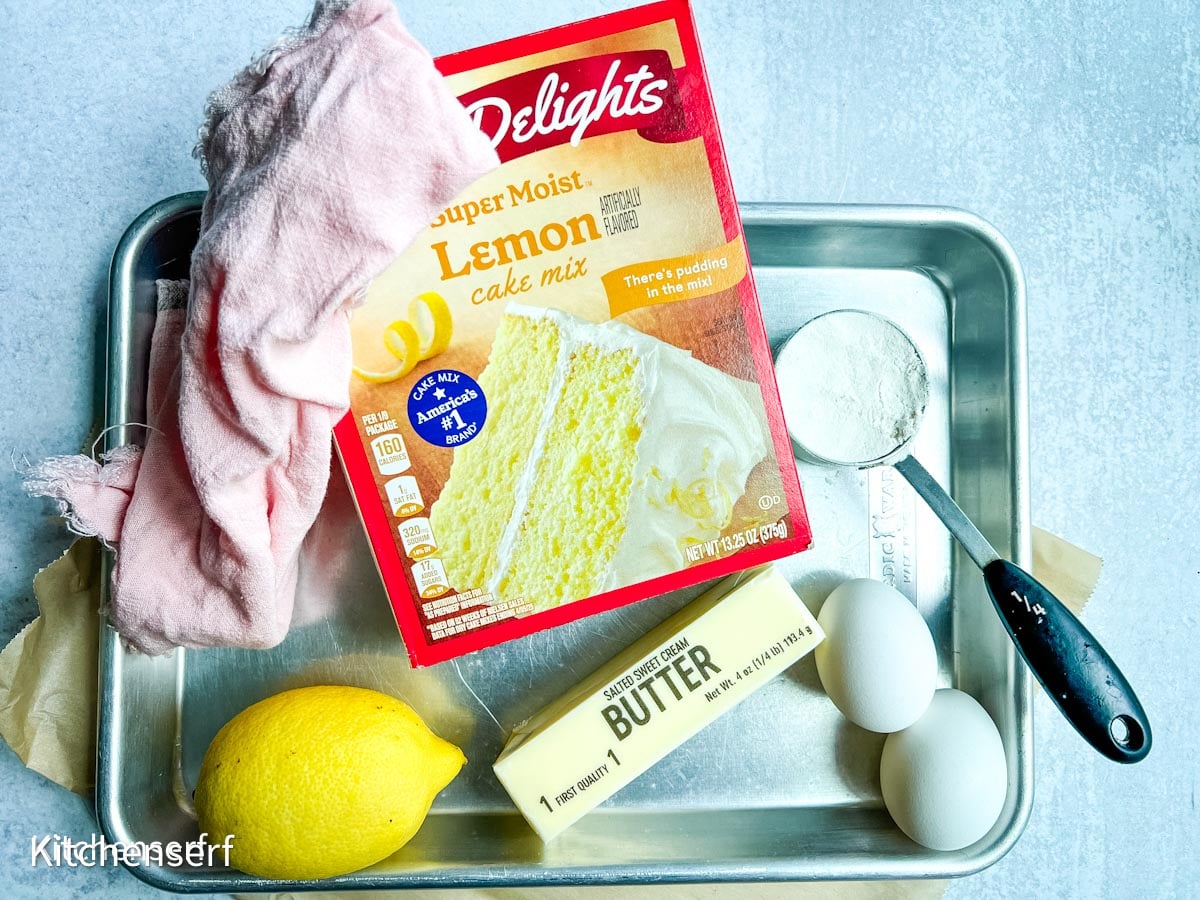 A box of lemon cake mix, butter, eggs, a lemon, flour, and a measuring spoon are arranged on a baking tray—everything you need to whip up lemon cake mix cookies.
