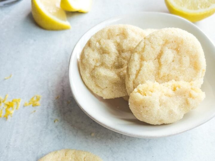 Three soft lemon sugar cookies in a white bowl with lemon wedges and zest scattered nearby on a light surface.