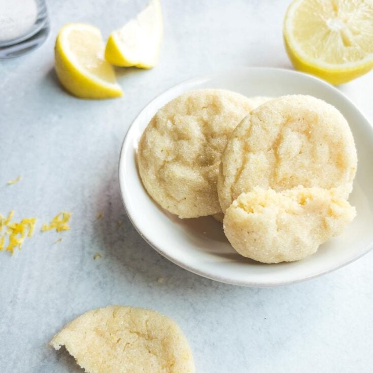 Three soft lemon sugar cookies in a white bowl with lemon wedges and zest scattered nearby on a light surface.