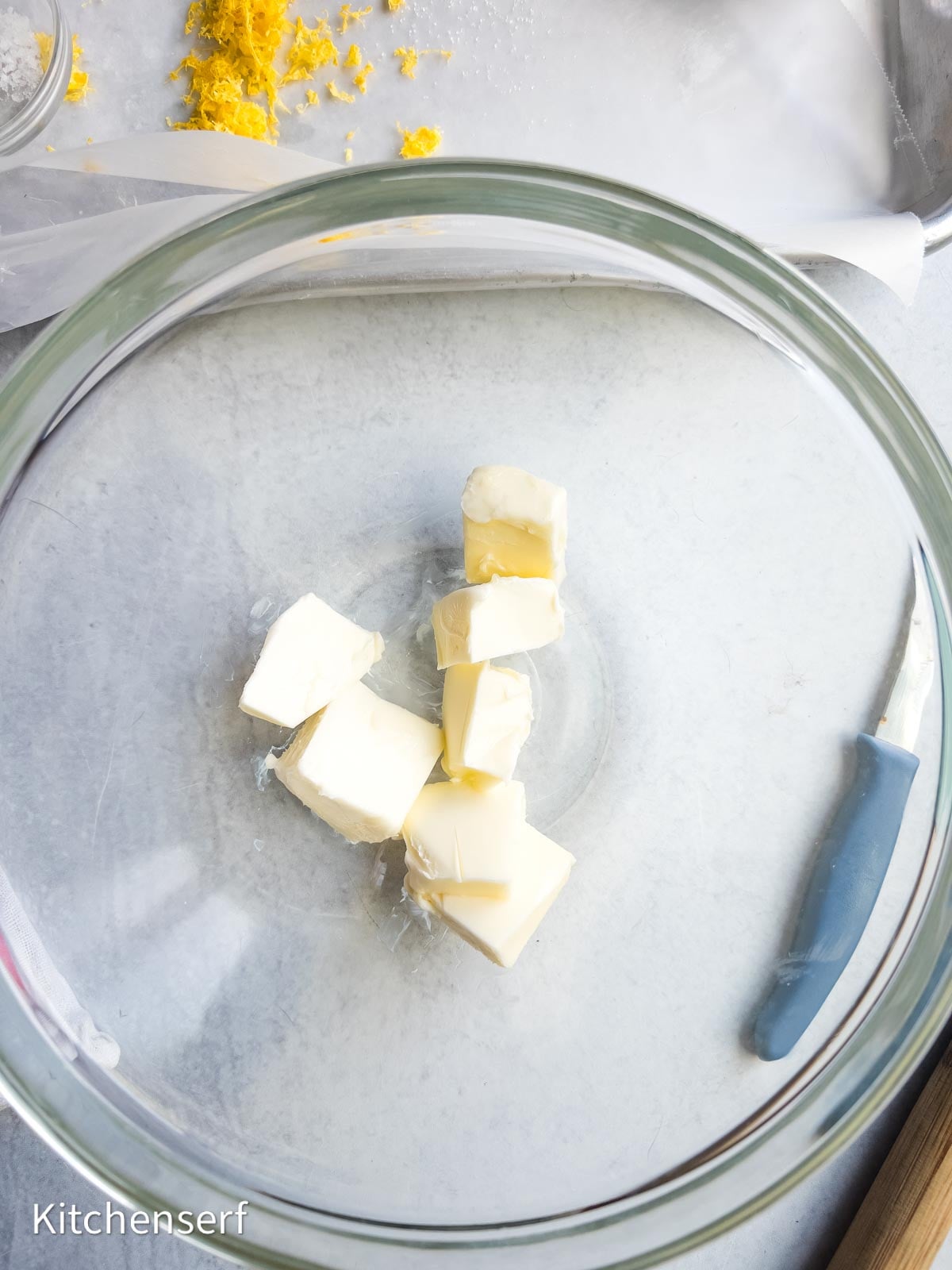 Cubed butter in a glass mixing bowl with a paring knife and lemon zest on a nearby tray, ready to be transformed into soft lemon sugar cookies.