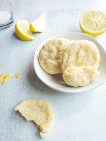 Three sugar cookies on a white plate with lemons nearby; one cookie has a bite taken out.