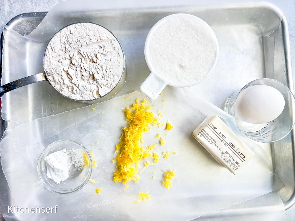 Flour, sugar, egg, butter, lemon zest, and baking powder arranged on a tray with wax paper underneath.