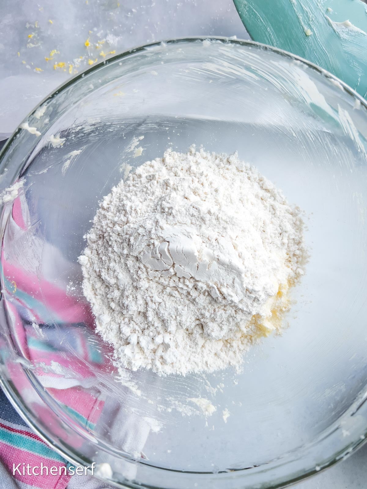 A glass bowl with flour and a bit of dough for soft lemon sugar cookies, on a countertop with a striped towel nearby.