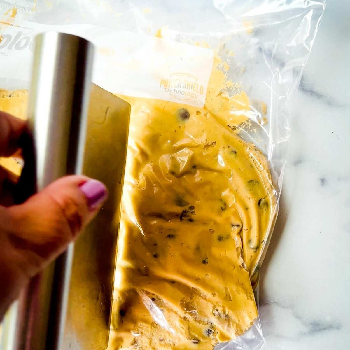 Hand holding a metal bench scraper to crush cookie dough in a plastic bag on a white surface—showing what to use instead of a cookie scoop.