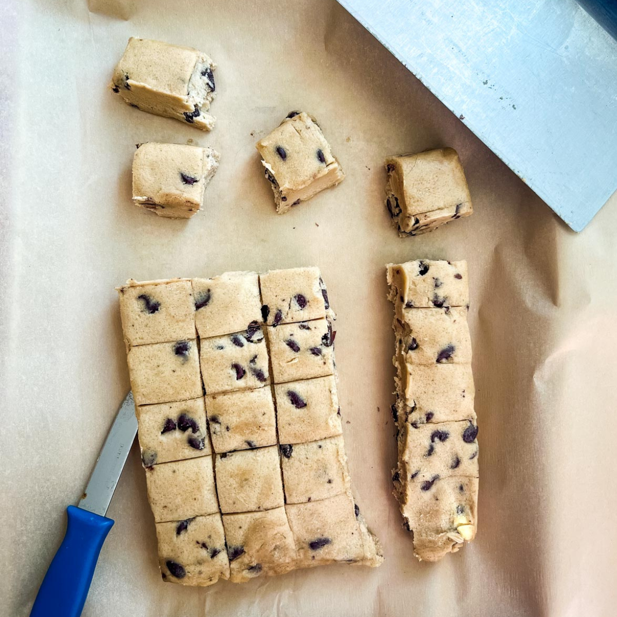 Squares of chocolate chip cookie dough being cut on parchment paper with a knife and bench scraper nearby—perfect options for what to use instead of a cookie scoop.