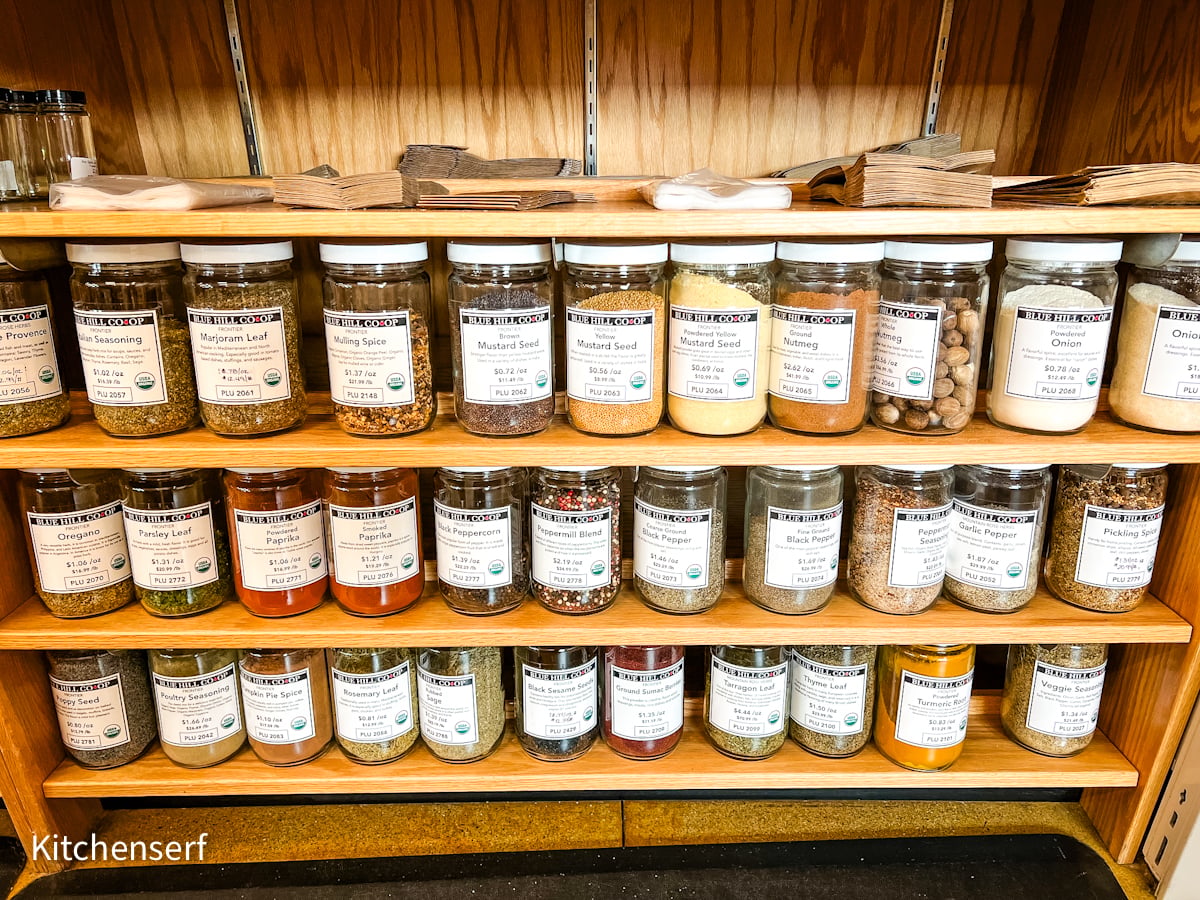 Glass jars of various spices and herbs are  arranged on store shelves.
