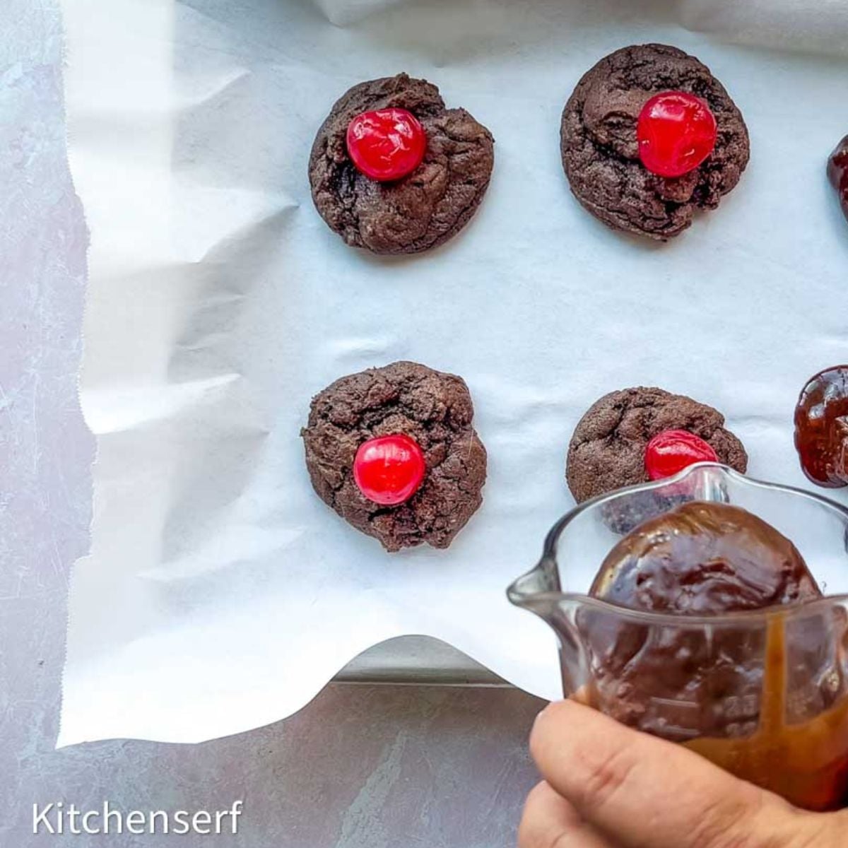 Hand spooning chocolate onto cookies topped with cherries on a parchment-lined baking sheet—perfect homemade cookies for gifts.