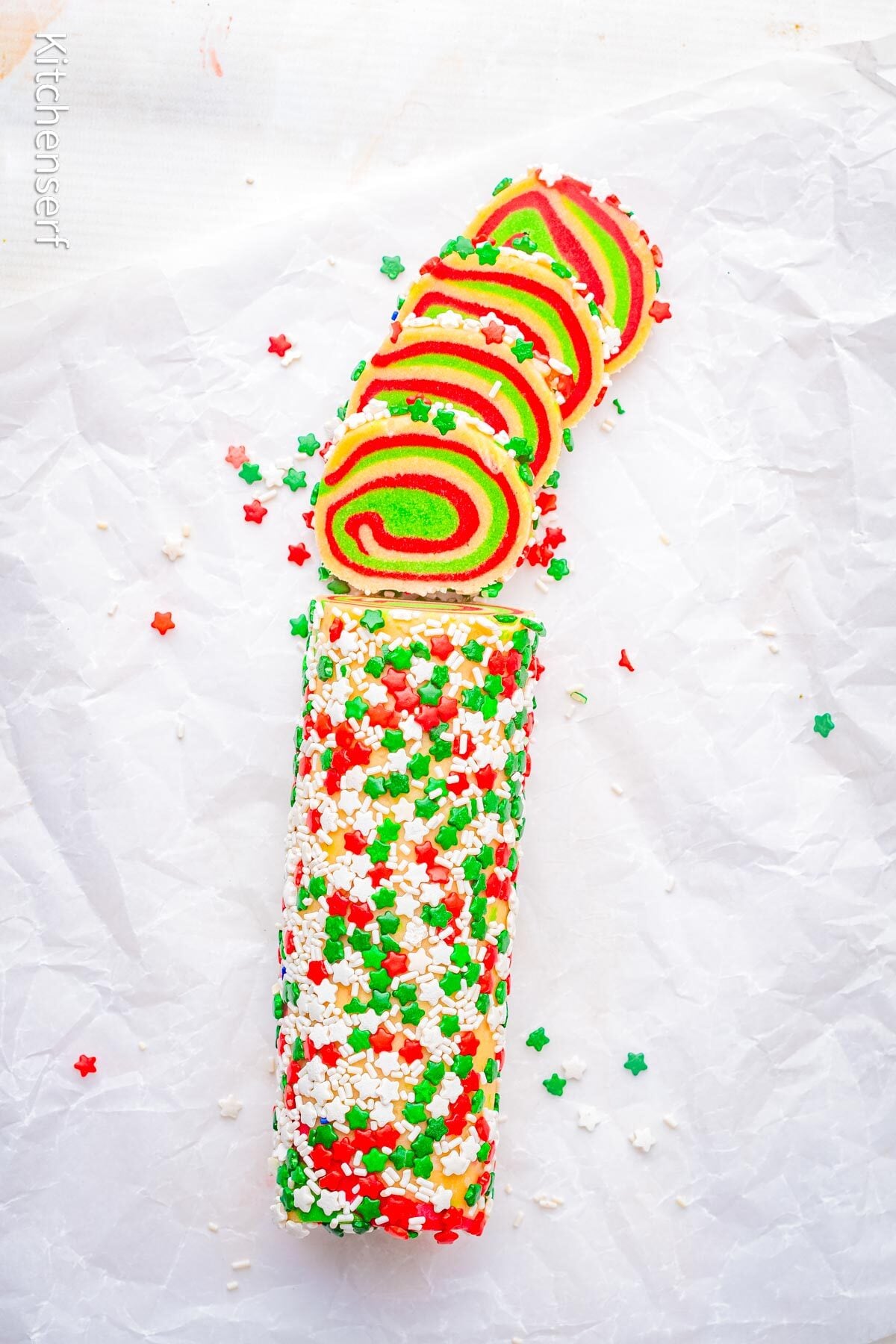 A sliced peppermint pinwheel cookie roll covered in red, green, and white star sprinkles rests on parchment paper.